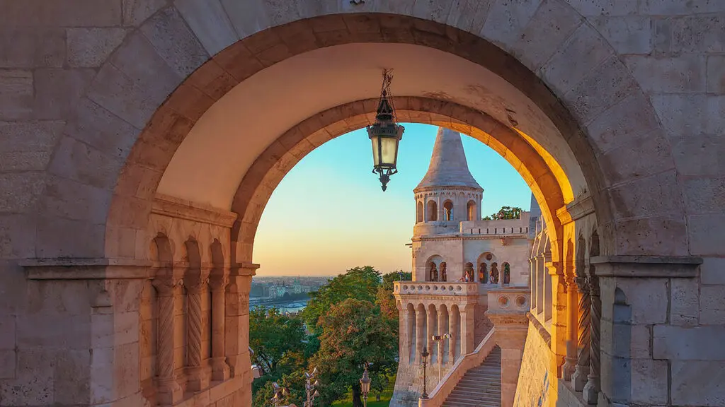 View through an archway of the Fisherman's Bastion at sunrise, showcasing its neo-Gothic and neo-Romanesque style against a backdrop of the cityscape and sky. One of the must-visit things to do in Budapest for breathtaking views.