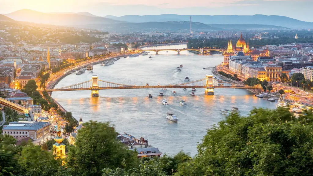 An aerial view of the Danube River at twilight showcases multiple illuminated bridges, including the Chain Bridge, and the Parliament Building in Budapest, Hungary. Among the top things to do in Budapest is admiring this breathtaking nighttime panorama.