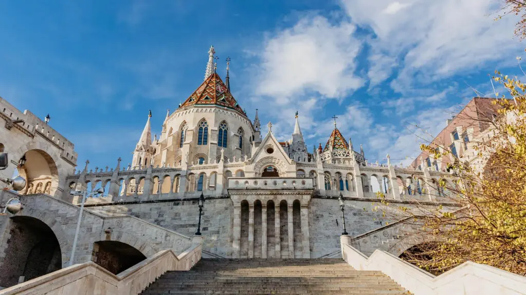 A grand building with ornate architecture features pointed towers and a colorful, tiled roof. It is framed by a clear, blue sky and flanked by steps and autumnal trees, making it one of the must-see sights for things to do in Budapest.