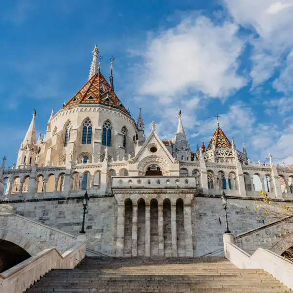 A grand building with ornate architecture features pointed towers and a colorful, tiled roof. It is framed by a clear, blue sky and flanked by steps and autumnal trees, making it one of the must-see sights for things to do in Budapest.