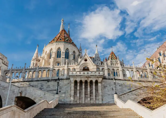 A grand building with ornate architecture features pointed towers and a colorful, tiled roof. It is framed by a clear, blue sky and flanked by steps and autumnal trees, making it one of the must-see sights for things to do in Budapest.