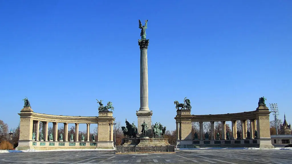 A monumental square featuring a high column with a statue on top, surrounded by semi-circular colonnades with statues, under a clear blue sky—one of the must-see things to do in Budapest.