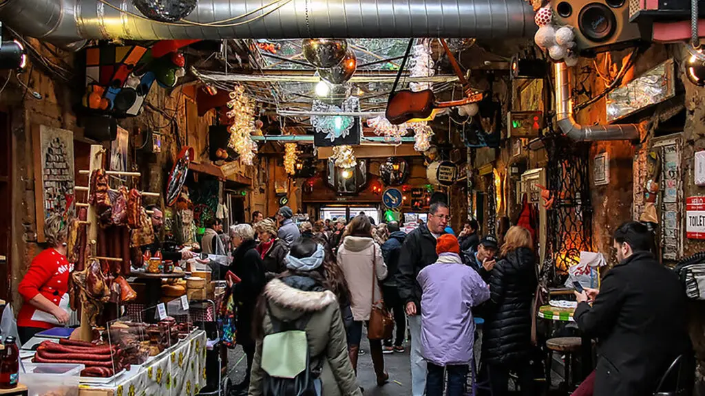 A bustling indoor market with various vendors and shoppers offers an eclectic array of things to do in Budapest. The space is adorned with hanging lights, pipes, and quirky decor, creating a lively atmosphere where people eagerly browse and purchase unique items.