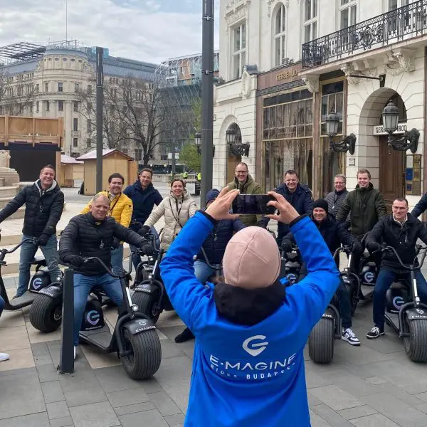 Emberek egy csoportja elektromos robogón ülve pózol egy szabadtéri városi környezetben, történelmi épületekkel és egy kőszoborral a háttérben, és élvezi a népszerű budapesti tenivalók karácsonykor. Egy kék kabátos személy fényképezi őket.