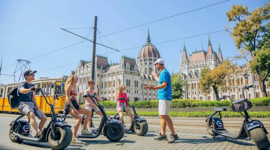 A group of four people on electric scooters listens to a guide in a blue shirt in front of a historic building with towers and a dome. A tram is passing by on the left.