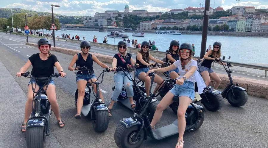 A group of eight women posing on electric scooters by a riverside path with a cityscape in the background. All are wearing helmets and casual summer attire.