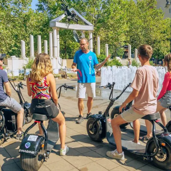 Egy kék inges idegenvezető négy emberhez beszél, akik elektromos robogón ülnek egy szökőkút és park közelében, fákkal és fehér pavilonnal a háttérben.