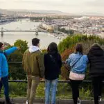 Five people stand at a railing overlooking a cityscape with a river and bridges. One person points towards the city. The weather is clear.