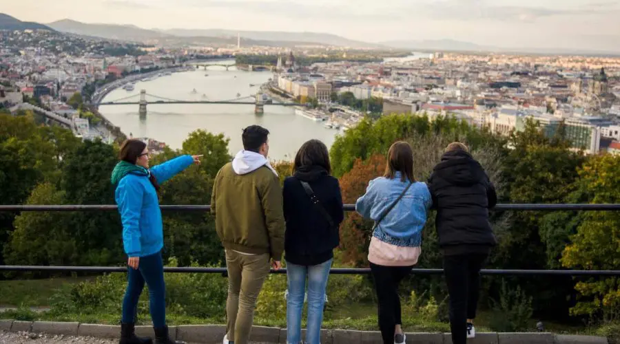 Five people stand at a railing overlooking a cityscape with a river and bridges. One person points towards the city. The weather is clear.