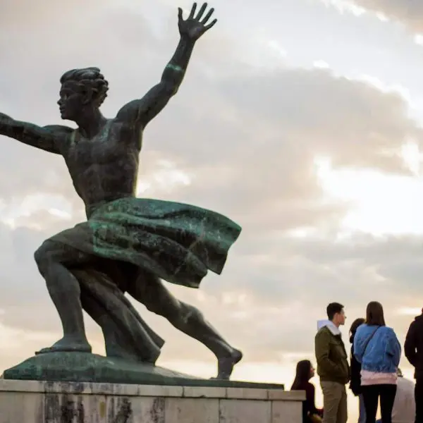 Bronze statue of a man with a palm frond, with a group of people standing near its base. Cloudy sky in the background.
