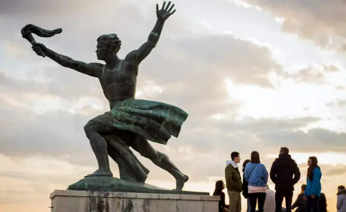 Bronze statue of a man with a palm frond, with a group of people standing near its base. Cloudy sky in the background.