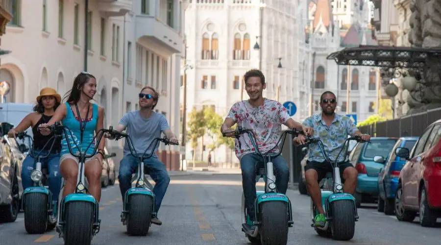 Four people are riding electric scooters down a city street, flanked by parked cars on both sides. The riders appear to be enjoying themselves on a sunny day.