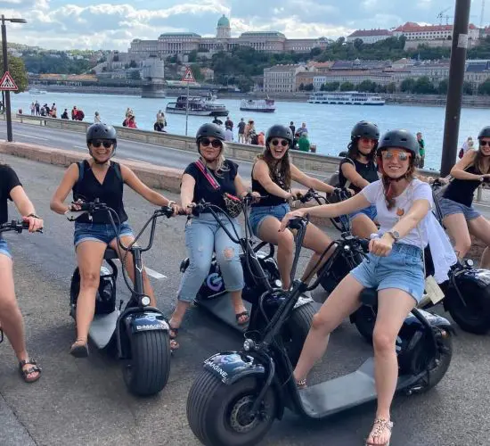 A group of eight people wearing helmets sit on electric mopeds near a waterfront with buildings and boats in the background.