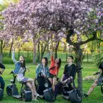 Five people on electric scooters gather under a blooming cherry blossom tree in a park, looking and pointing up at the branches.