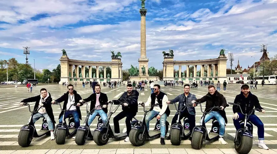 A group of seven people on motorized scooters pose in front of a large historic monument and colonnades in an open plaza under a clear sky.