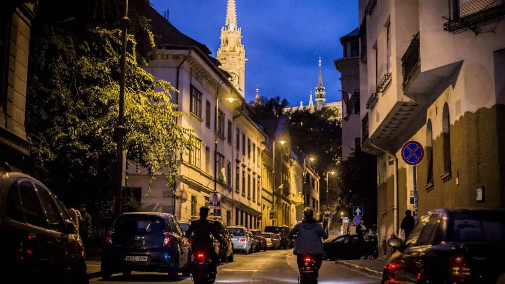 Three people on motorcycles ride through a narrow street lined with cars, with tall, illuminated church spires in the background at dusk.