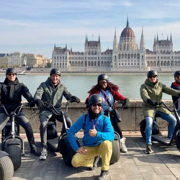 A group of people wearing helmets pose on electric scooters in front of a large historic building by a river.