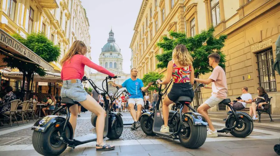 Four people on electric scooters stop on a city street between buildings. Two women and two men are facing each other, with a domed structure visible in the background.