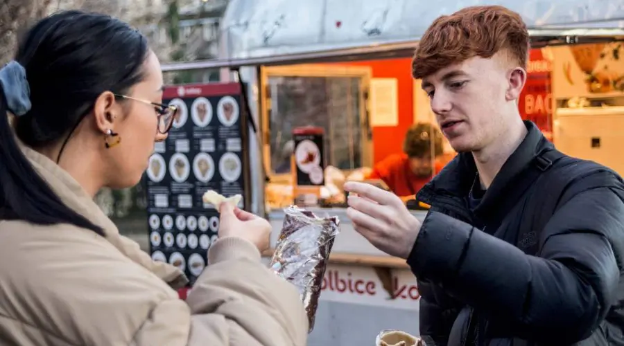 Két ember egy élelmiszerszállító teherautó közelében áll, és ételt osztanak. A bal oldali személy pakolást tart, a jobb oldali pedig falatot kínál. A háttérben egy menü látható.