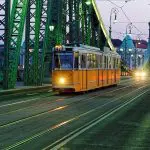 An orange tram travels across a green bridge at dusk, with cars following behind and buildings visible in the background.