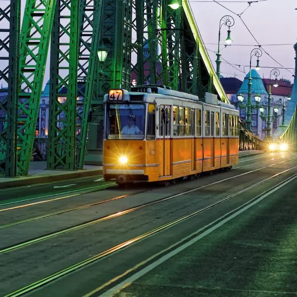 An orange tram travels across a green bridge at dusk, with cars following behind and buildings visible in the background.