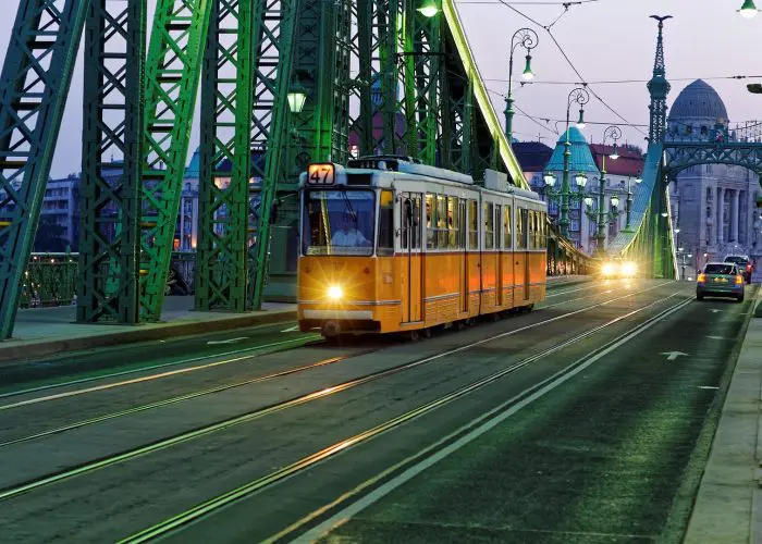 An orange tram travels across a green bridge at dusk, with cars following behind and buildings visible in the background.