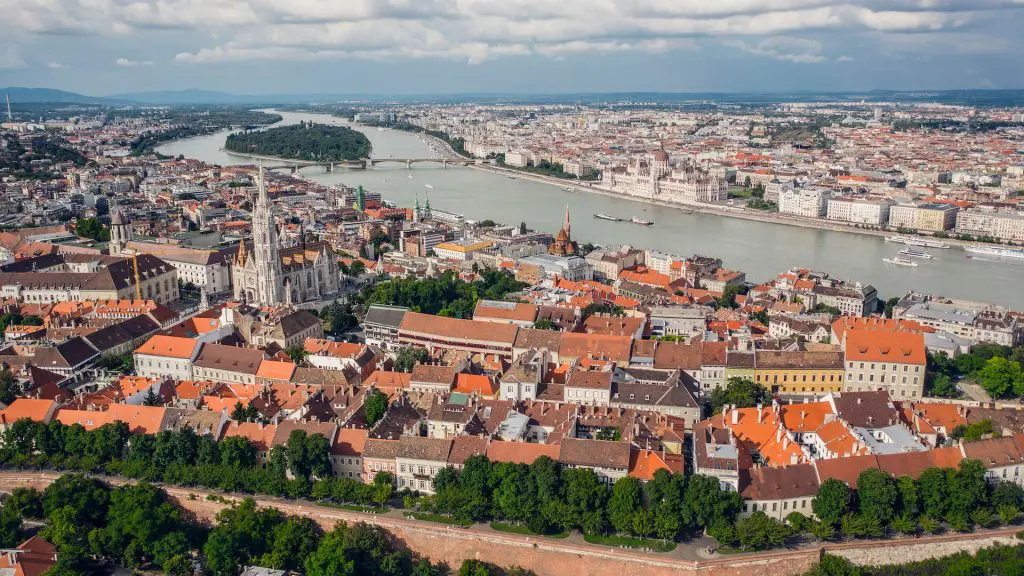 Aerial view of Budapest showcasing the Danube River, the Parliament building on the river bank, and the city's historic buildings and rooftops with scattered greenery.