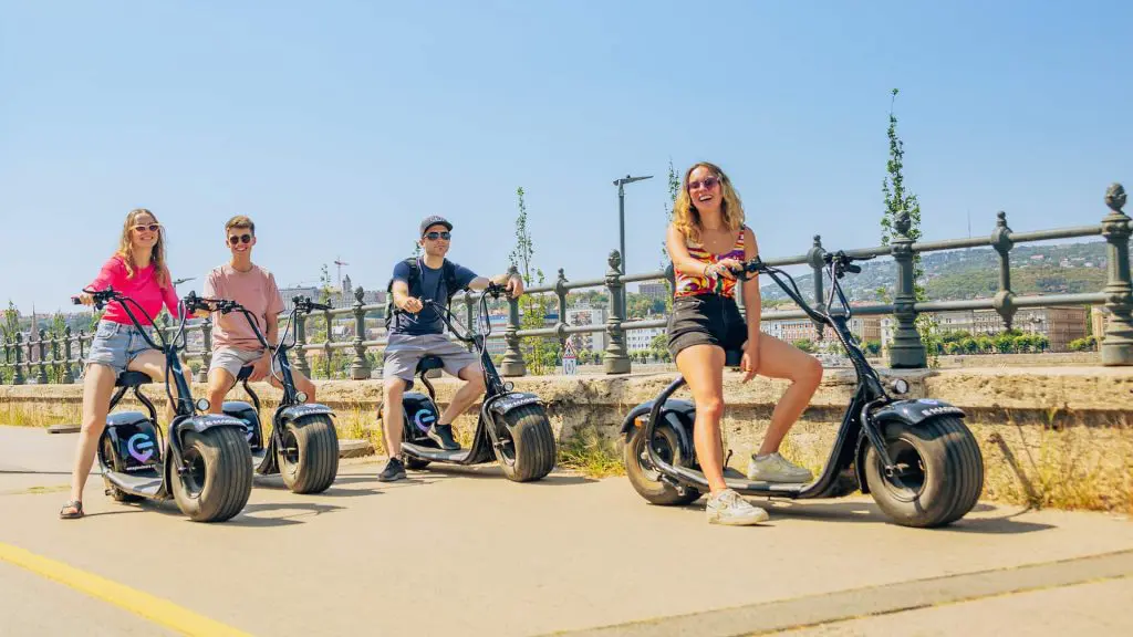 Four people riding electric scooters on a sunny day near a waterfront, smiling and wearing casual summer clothes.