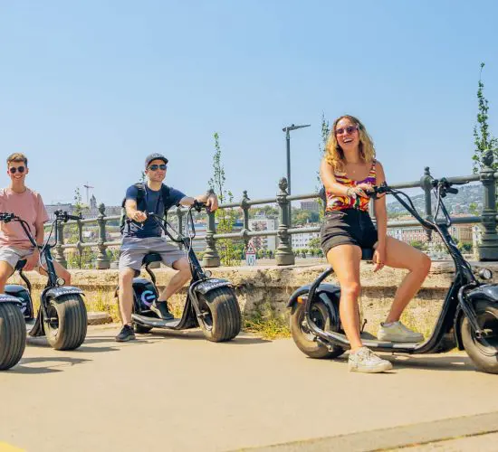 Four people riding electric scooters on a sunny day near a waterfront, smiling and wearing casual summer clothes.