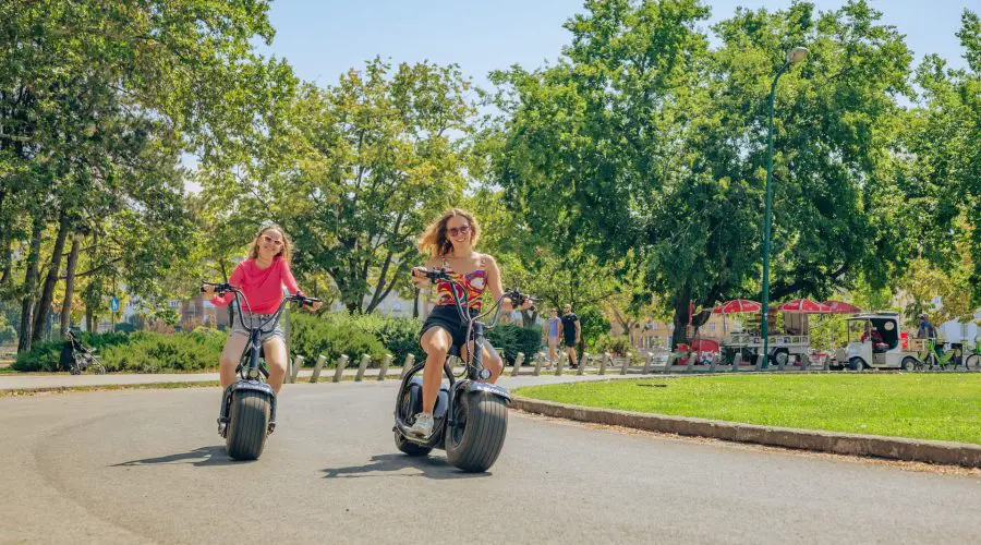 Two women ride electric scooters on a paved path in a sunny park, with trees and people in the background.