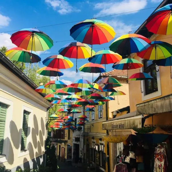 A narrow street is decorated with hanging multicolored umbrellas. The street is lined with shops and buildings, and there is a blue sky visible above.