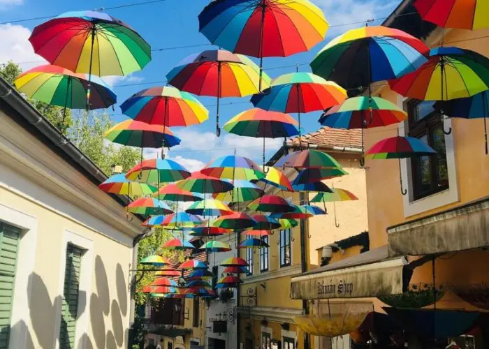 A narrow street is decorated with hanging multicolored umbrellas. The street is lined with shops and buildings, and there is a blue sky visible above.