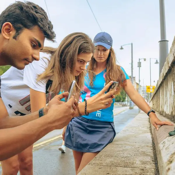 Three people are intently looking at and photographing small green sculptures on a concrete wall in an outdoor urban area.