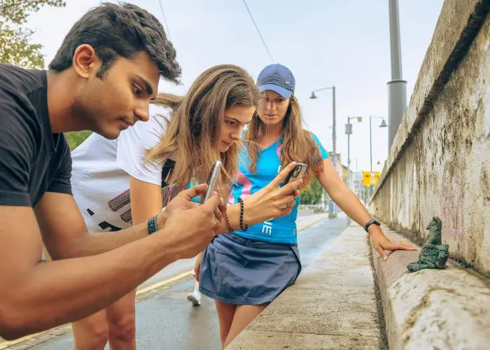 Three people are intently looking at and photographing small green sculptures on a concrete wall in an outdoor urban area.