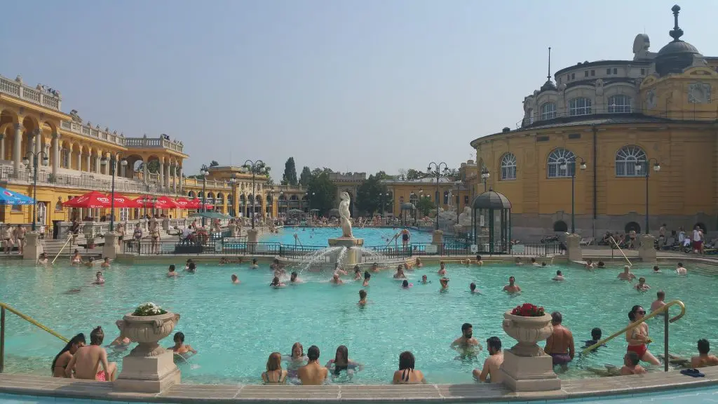 People swimming in an outdoor thermal pool surrounded by yellow historic buildings on a sunny day, just a short stroll from the significance of Heroes’ Square.