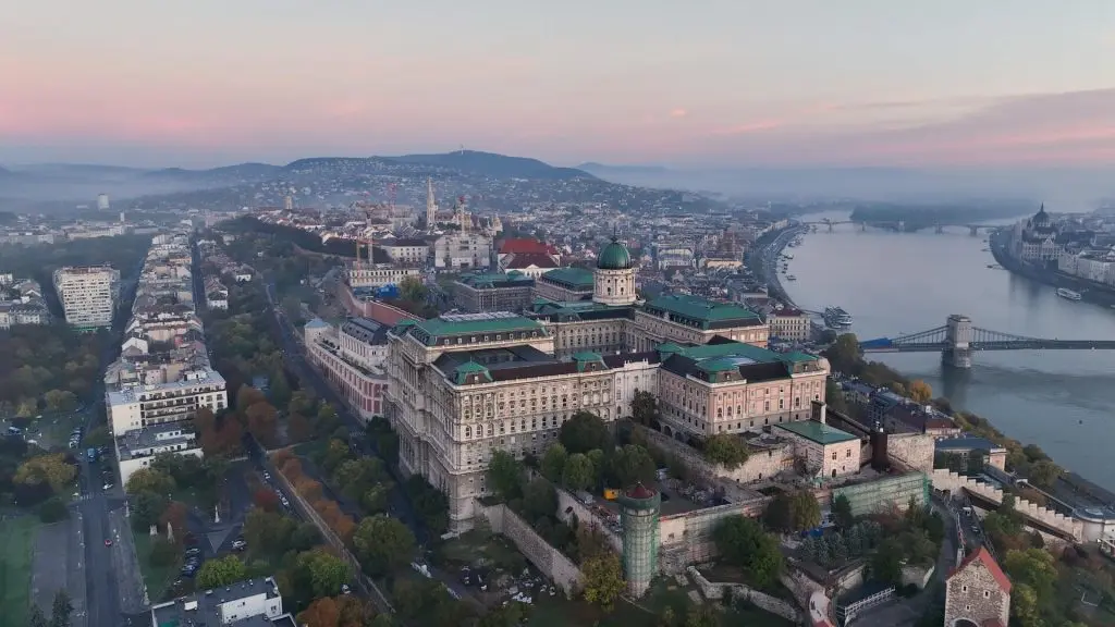 Aerial view of Buda Castle in Budapest, Hungary, with the Danube River on the right and the cityscape extending into the distance under a pink and blue sky, revealing the fascinating history of Buda Castle Budapest.