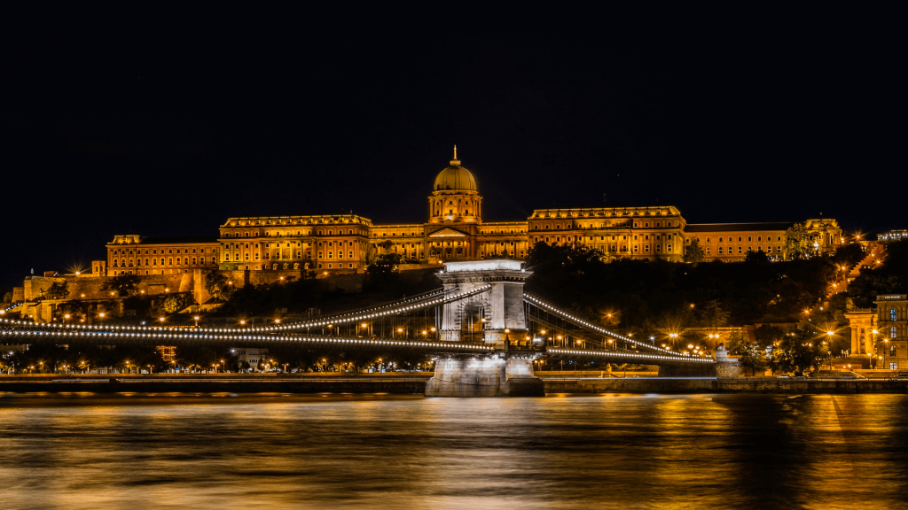 Night view of a brightly illuminated historic building and a suspended bridge over a river with reflections of the lights on the water surface – perfect for Budapest stag do ideas.