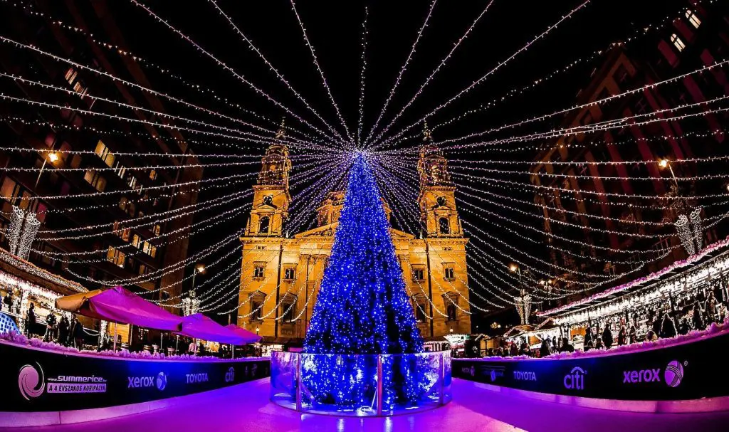 A large, illuminated Christmas tree with blue lights stands at the center of a decorated urban square during the night. Surrounding buildings and market stalls, reminiscent of the Budapest Christmas Market, are adorned with string lights.