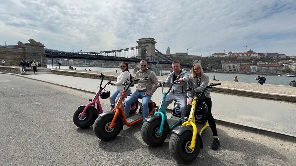 Four people are sitting on colorful electric scooters in front of a large bridge and river on a partly cloudy day, offering a glimpse into what is the best way to explore Budapest.