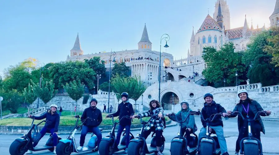 A group of seven people on electric scooters pose in front of a historic building with castle-like architecture and a church tower in the background, showcasing the charm of Budapest Scooter Tours.