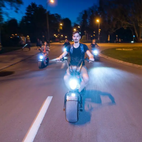 A person rides an electric scooter on a street at night, followed by others on similar scooters. Streetlights illuminate their path, revealing trees in the background. It's a scene straight out of Budapest Scooter Tours, capturing the essence of nocturnal adventure.