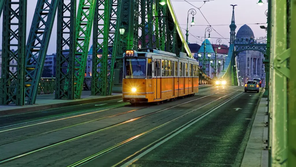 A yellow tram travels across a green bridge at dusk, with city buildings visible in the background.