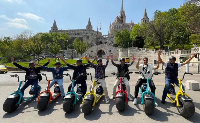 A group of eight people on electric scooters raise their arms in front of a historic building with multiple towers and a staircase, showcasing the joy of discovering where and how to rent an electric scooter in Budapest.