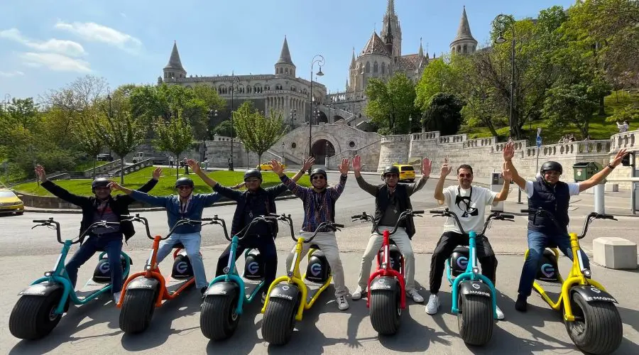 A group of eight people on electric scooters raise their arms in front of a historic building with multiple towers and a staircase, showcasing the joy of discovering where and how to rent an electric scooter in Budapest.