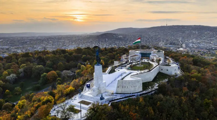 Aerial view of the Citadella fortress on Gellért Hill in Budapest, Hungary, with the Liberty Statue and large Hungarian flag, surrounded by autumn trees at sunset—perfect for a memorable Budapest citadel tour.