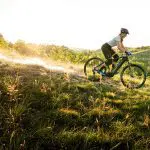 A person rides a mountain bike down a grassy hill during sunset.