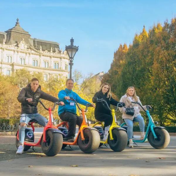 Four people riding colorful electric scooters are lined up in a park, part of the All In Budapest Tour, with an ornate building and trees in the background. The sunny day highlights the autumn foliage.