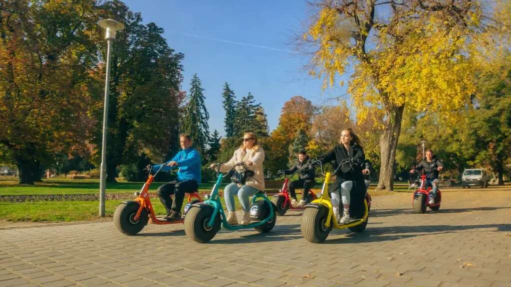 Five people glide on colorful electric scooters through a sunny park, part of the All In Budapest Tour. They travel along a paved path bordered by autumn trees with vibrant yellow and green leaves under a clear blue sky.