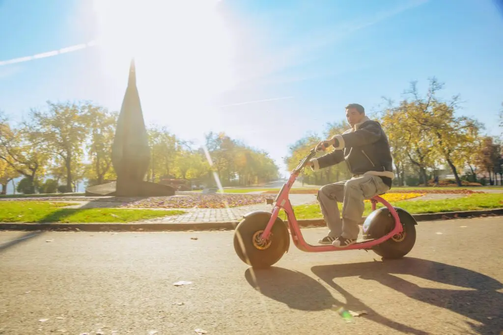 A person enjoys riding a pink electric scooter on a sunny day in the park in Budapest. Trees with autumn foliage provide a picturesque backdrop, and the path is beautifully illuminated by bright sunlight.