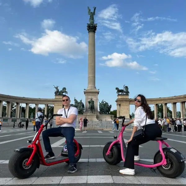 Two people riding e-scooters pose in front of the grand Heroes' Square monument.